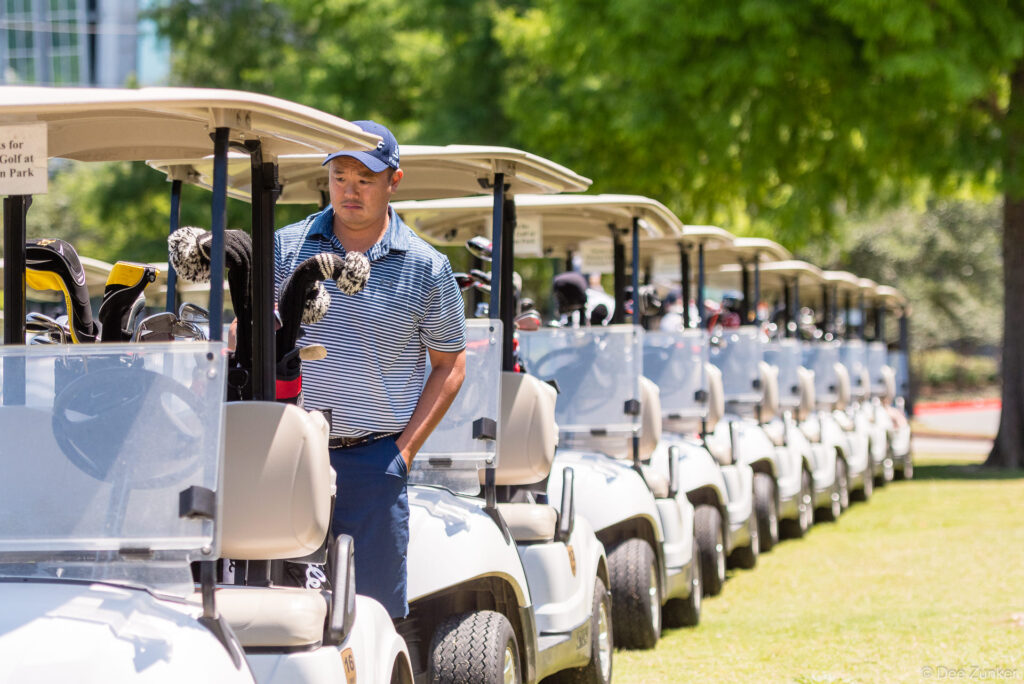 Golfer in striped polo and cap stands beside a long row of golf carts at Houston Metro Chamber of Commerce Golf Classic tournament.