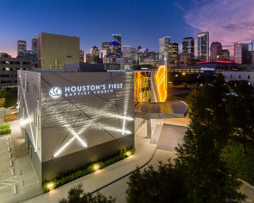 Aerial twilight exterior of Houston's First Baptist Church downtown campus with illuminated facade and Houston skyline in the background.