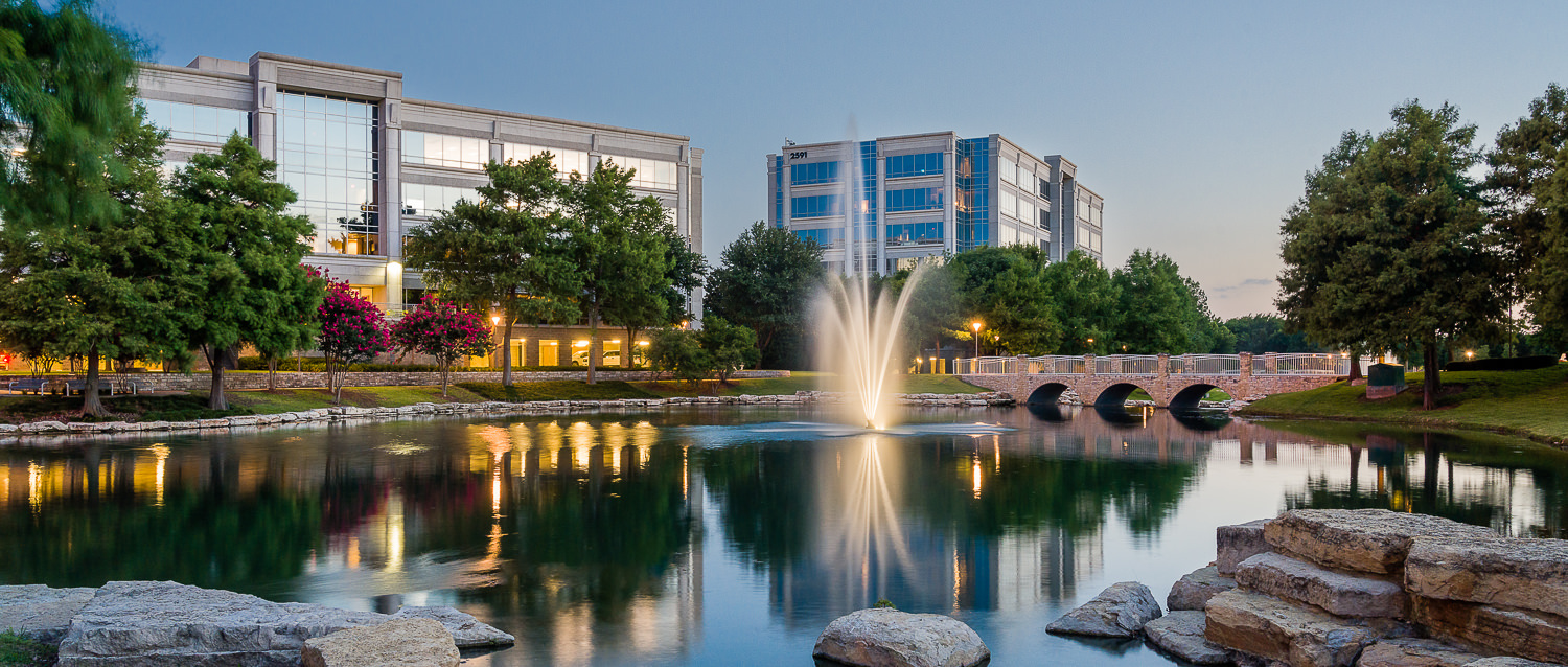 Architectural Landscape photography of Hall Park in Frisco TX at Twilight with lake, rocks, and fountain in front of office buildings 