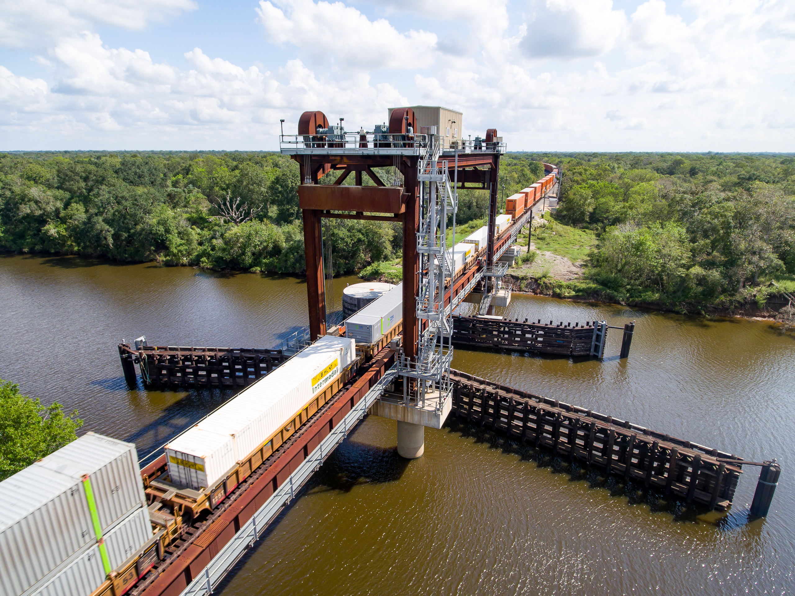Elevated drone photo of a freight train crossing the San Bernard River. The industrial vertical-lift bridge is seen from directly above.