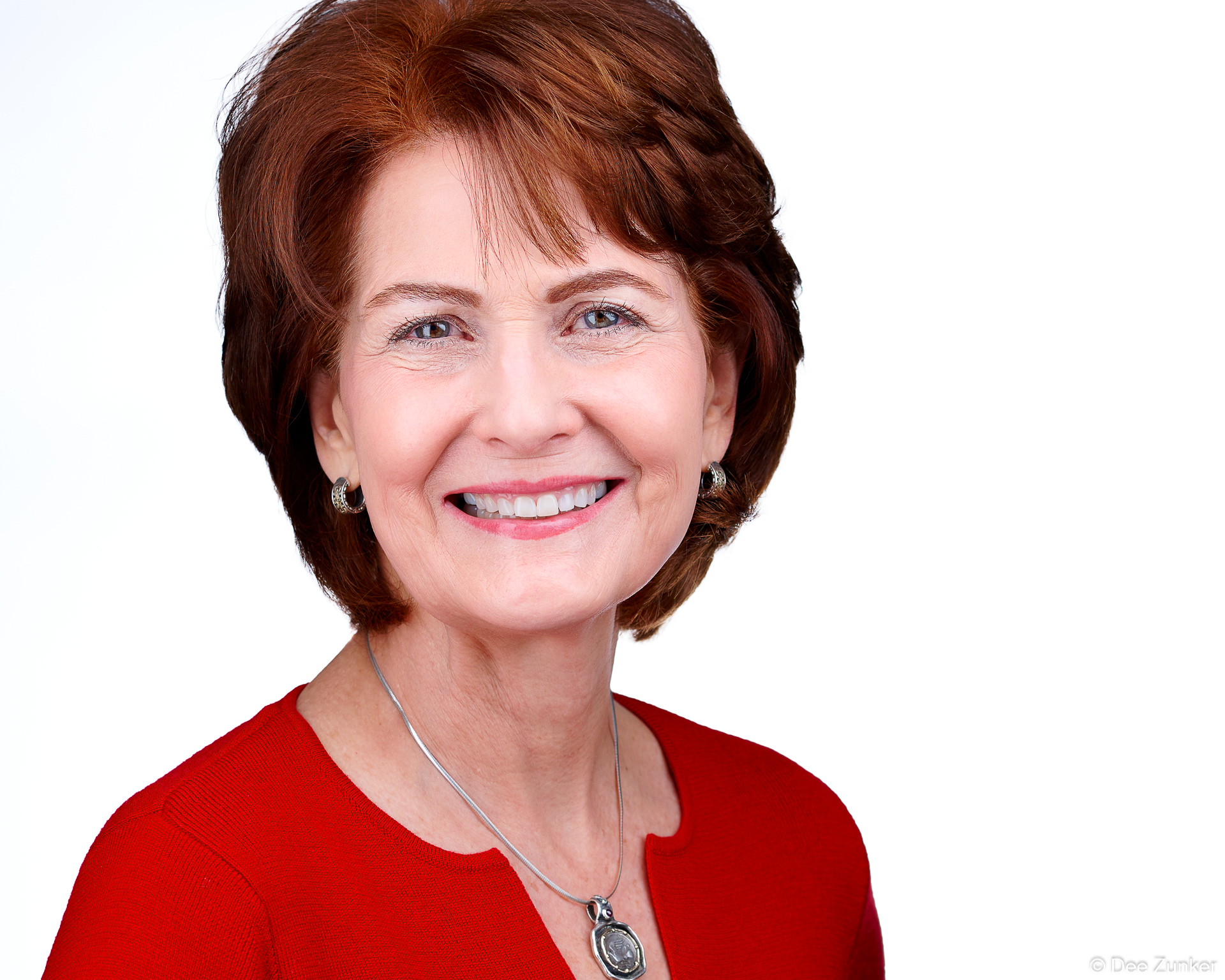 Smiling professional woman in red top, studio headshot with white backdrop, short auburn hair, silver necklace, Houston portrait session.