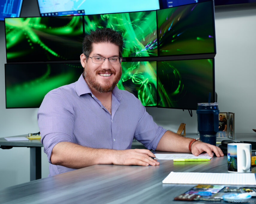 Smiling male professional in blue shirt seated at standing desk with six green-lit monitors, corporate headshot session, Houston TX.