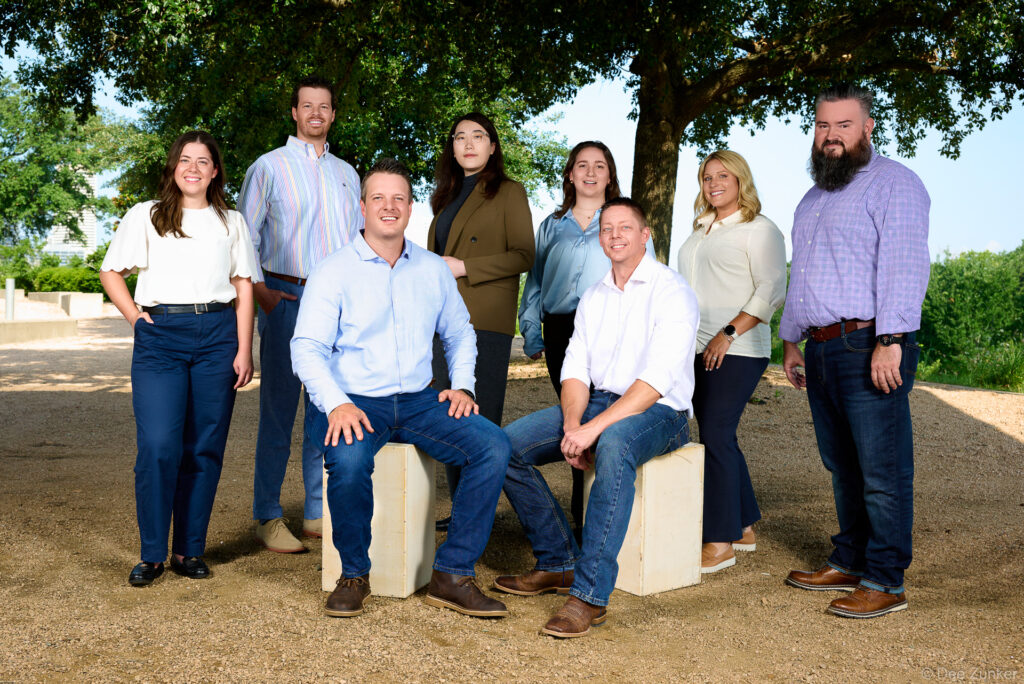 Nine corporate team members posed outdoors under oak trees on a gravel path for a Houston company group portrait session.