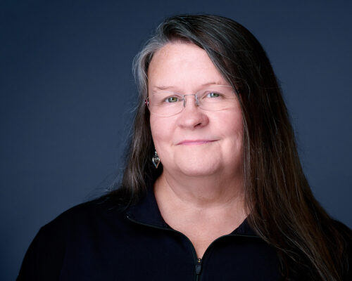 Professional Headshot of Dee Zunker on a dark blue background and wearing a black shirt.  