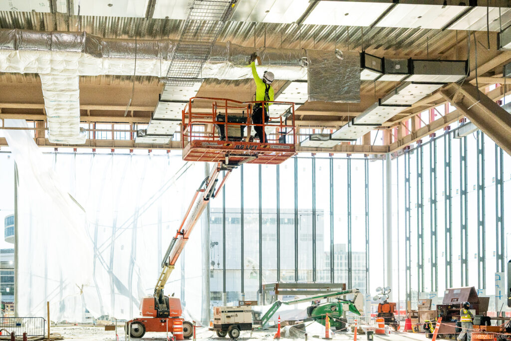 Construction worker on scissor lift installs insulated ductwork overhead at IAH Terminal B during active MEP systems progress documentation.