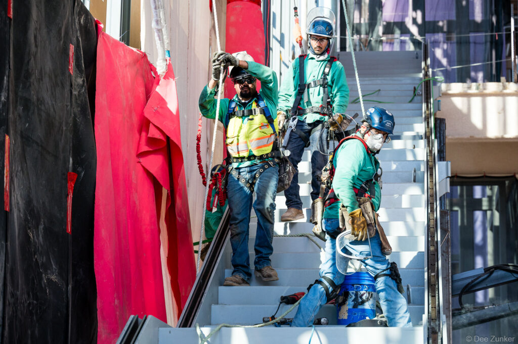 Three construction workers in safety harnesses and hard hats on exterior stairs at IAH Terminal B during active jobsite progress documentation.