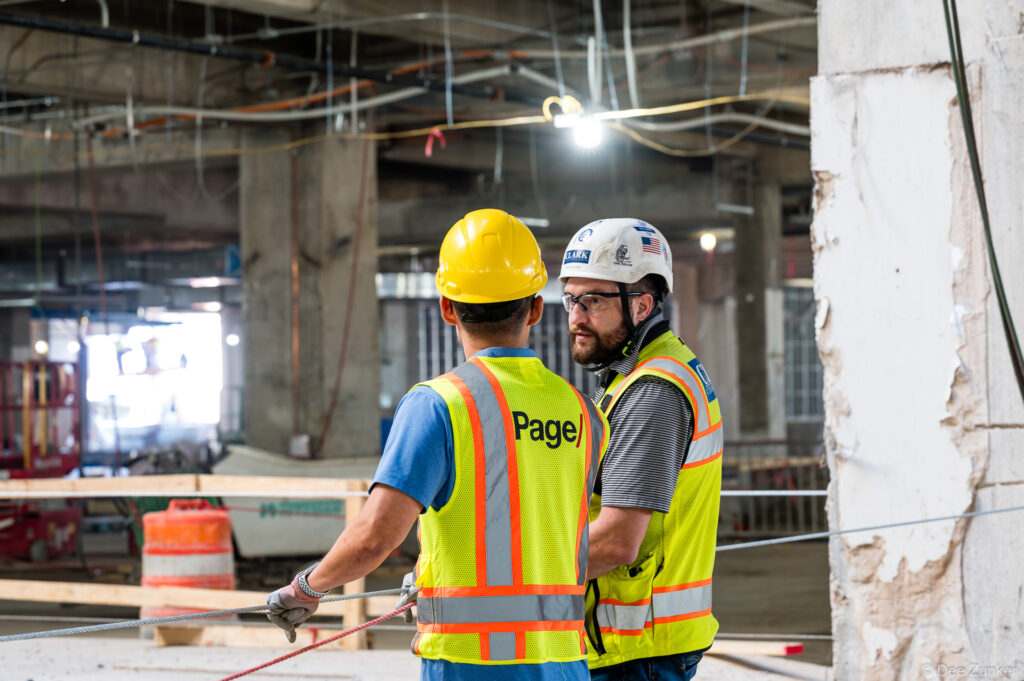 Two construction workers in high-visibility vests confer on the active jobsite interior of IAH Terminal B during structural progress work in Houston.