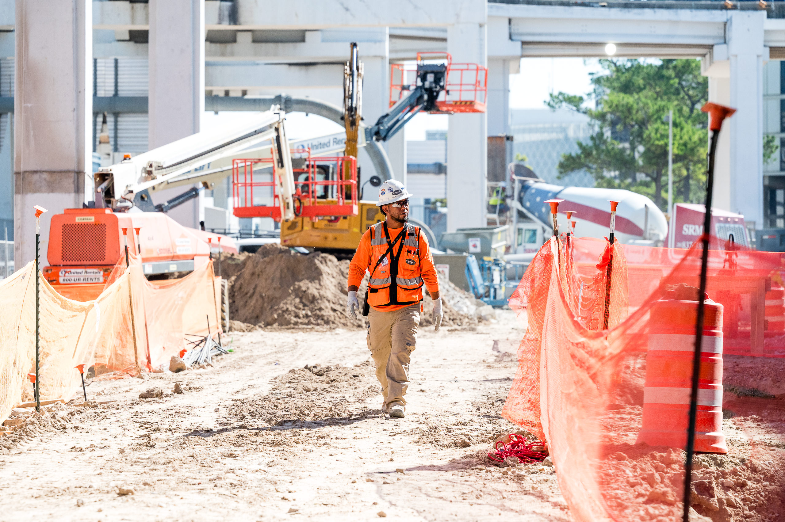 Construction worker walks through the site with orange barricades with equipment in teh backgrounds. 