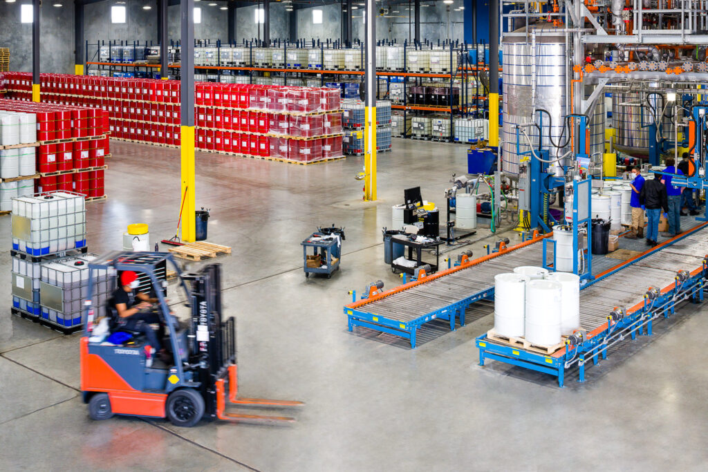 Industrial photo of a forklift in a busy warehouse. It's moving towards a production line, with stacked drums visible in the background.