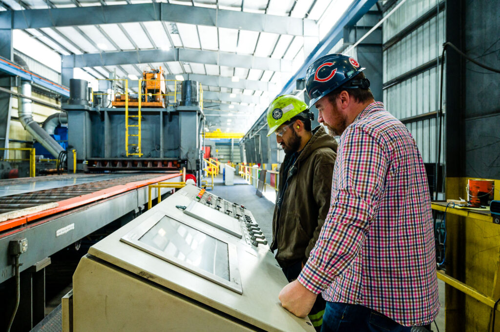 Industrial photo of two workers monitoring a computer console, managing a high-tech sheet metal assembly line in a factory.