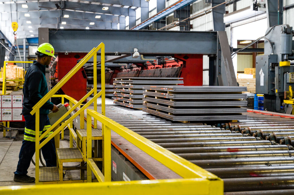 Industrial photo of a worker at a control panel. They are monitoring stacked metal plates rolling down a sheet metal conveyor line.