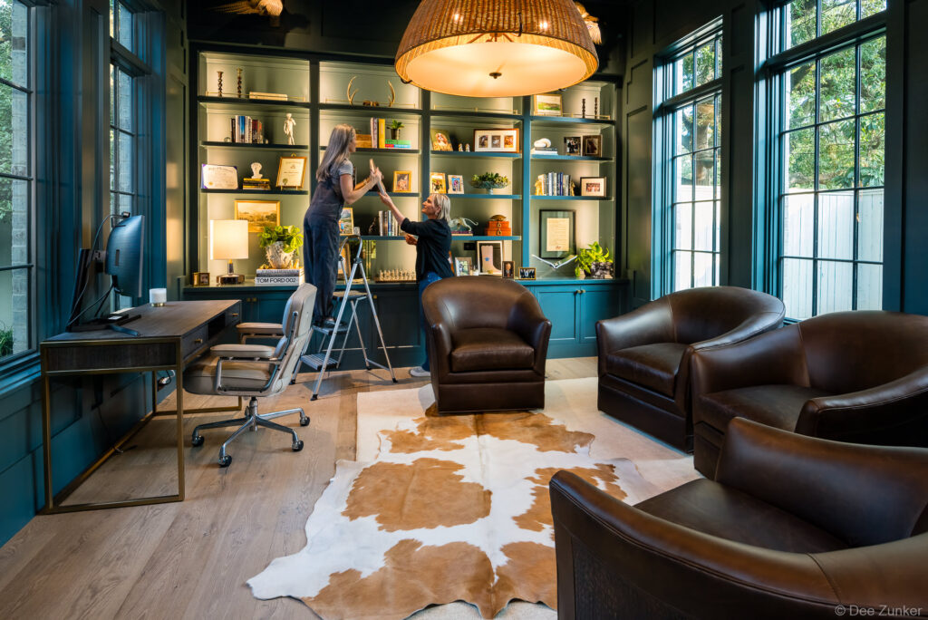 Two women arranging shelves in a dark teal home office library with leather chairs, cowhide rug, and built-in cabinetry, Houston residential interior.