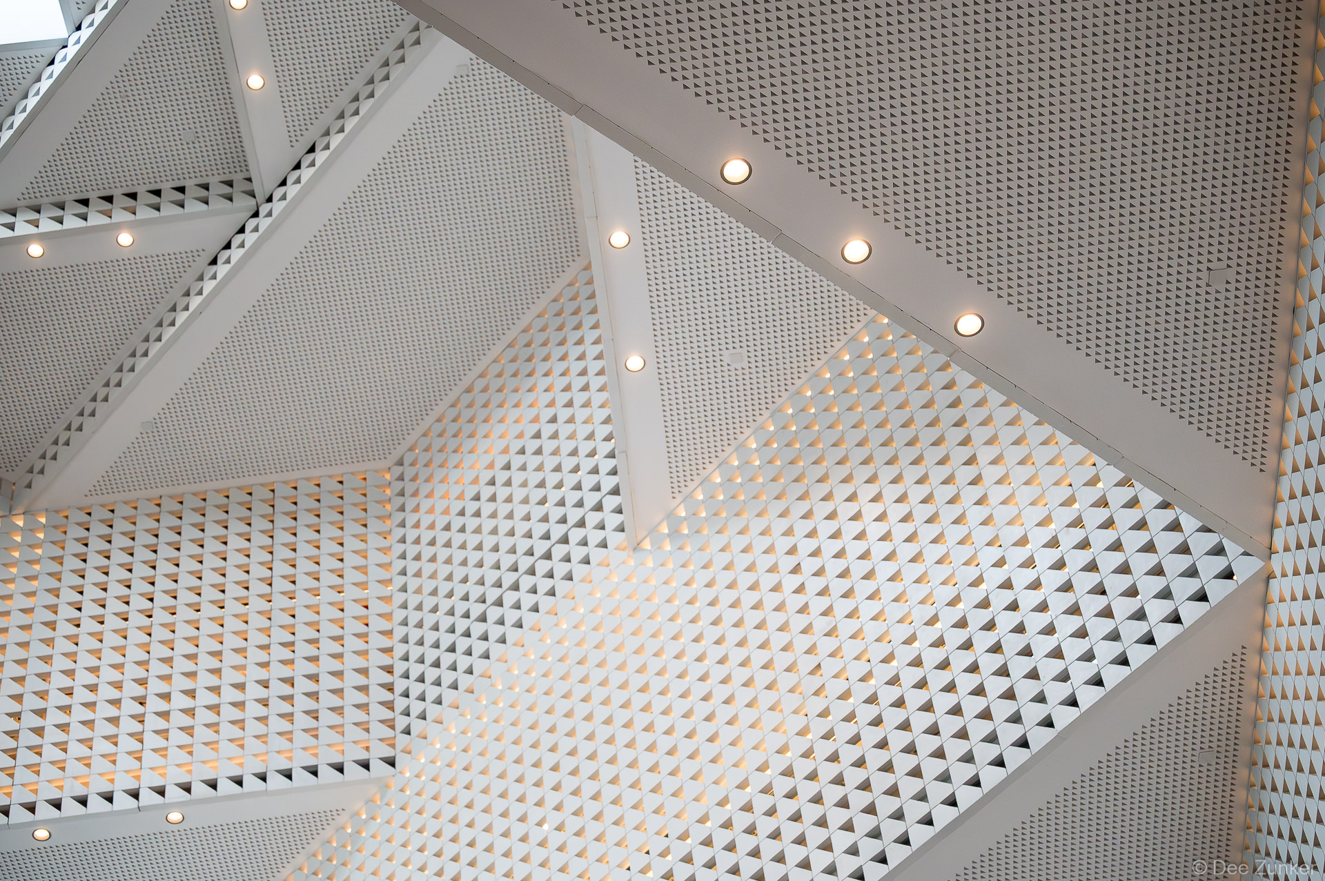 Upward view of the Ismaili Center Houston's triangular perforated white ceiling panels with structural beams and warm recessed lighting.