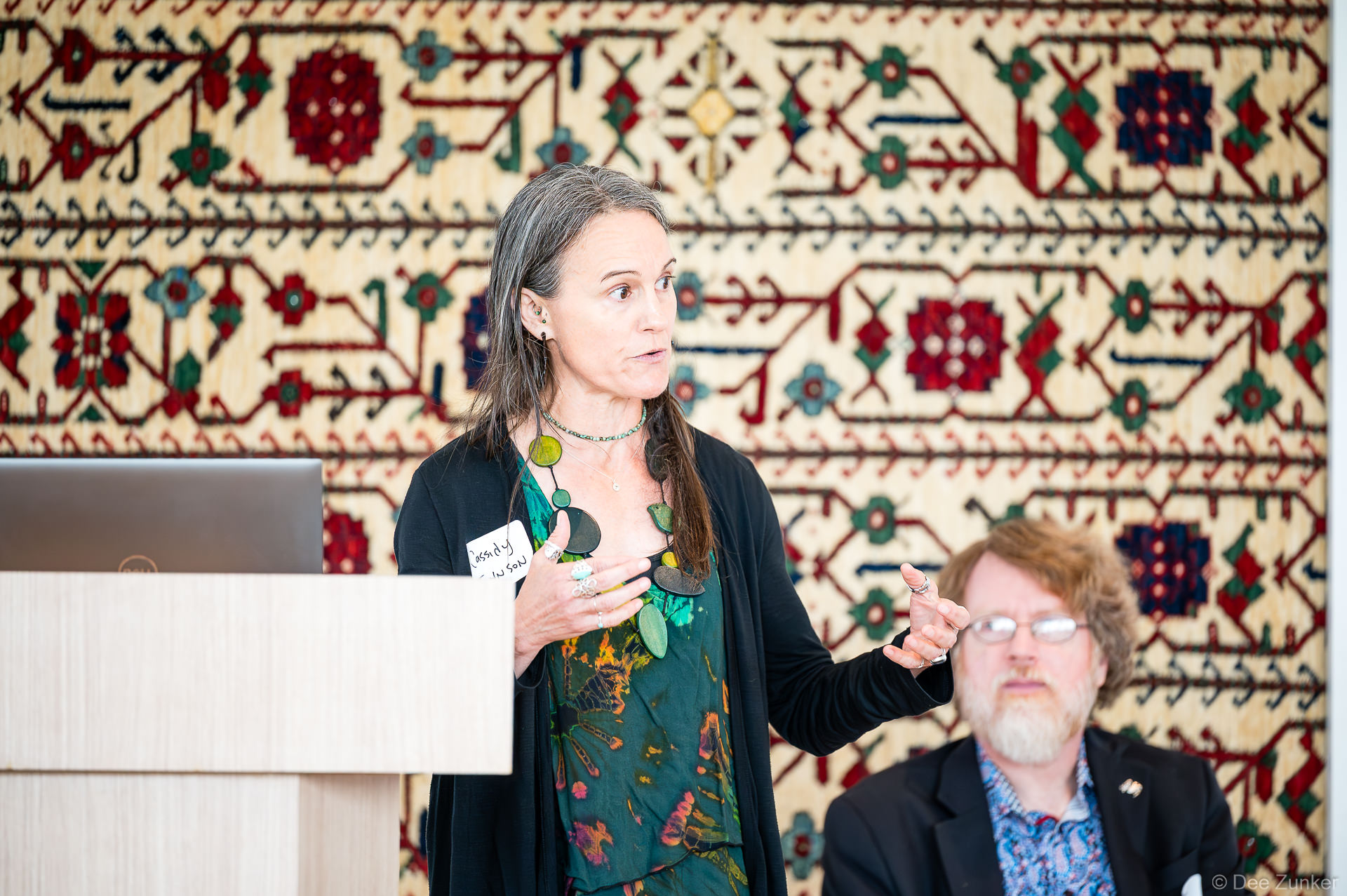 Dr. Cassidy Johnson speaker gestures at a podium during a panel discussion at Gulf Coast Green 2026, Ismaili Center Houston, ornate tapestry behind her.