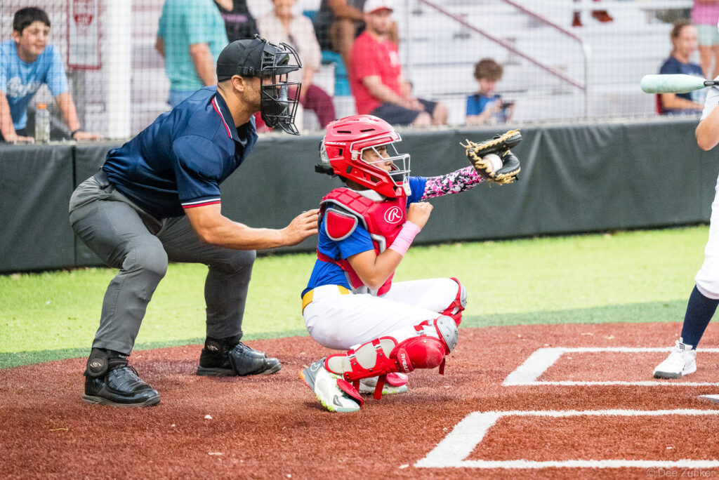Young catcher in red Rawlings gear receives a pitch at home plate with umpire behind, Bellaire Little League Mariners vs Twins game, Weston Field TX.