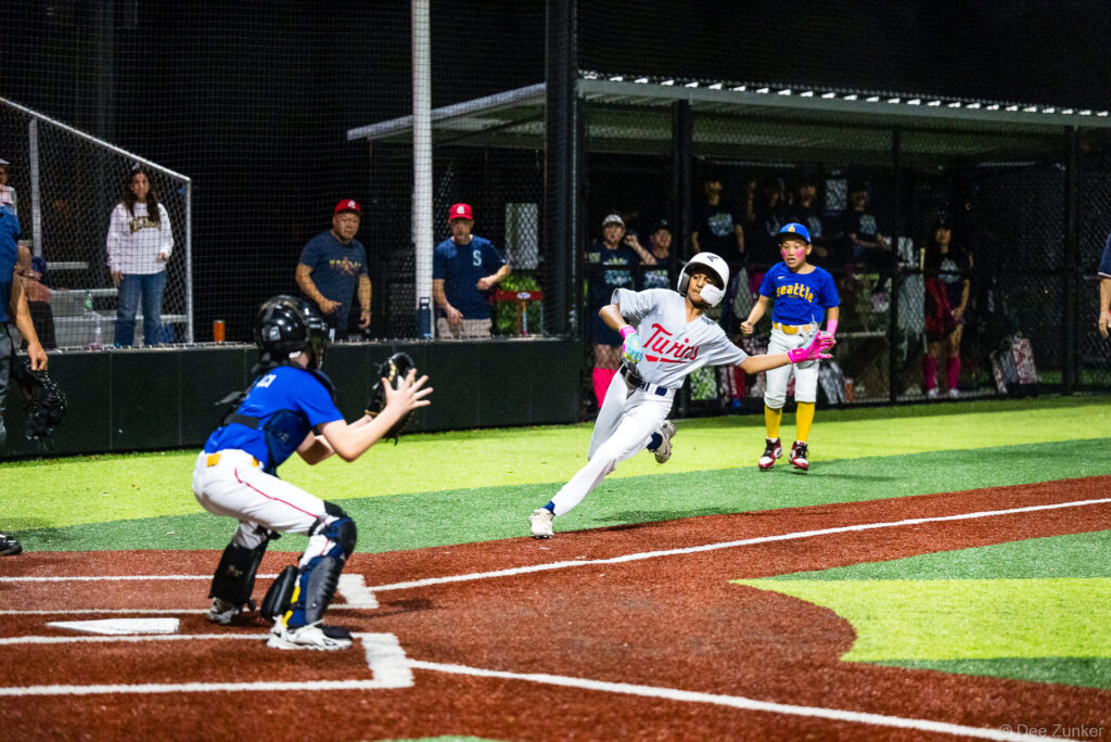 Twins baserunner rounds third and charges home as Mariners catcher prepares to receive the throw during a Bellaire Little League American League game at Weston or Tiras Field in Bellaire TX.