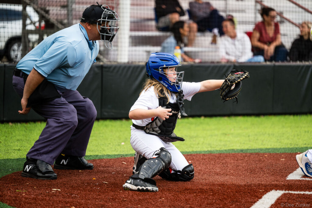 Young female catcher in blue helmet and full gear frames a target at home plate during a Rays vs Orioles Bellaire Little League game at Weston or Tiras Field in Bellaire TX.