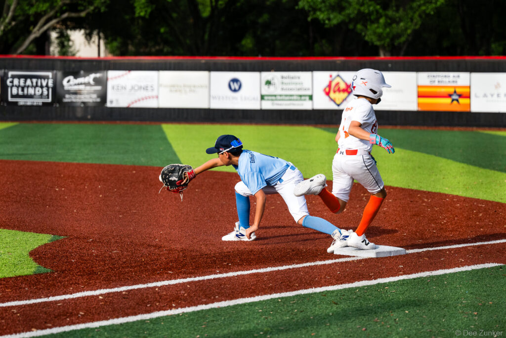 Young first baseman in blue stretches to catch ball as Orioles baserunner reaches first base at Bellaire Little League, Weston Field TX.