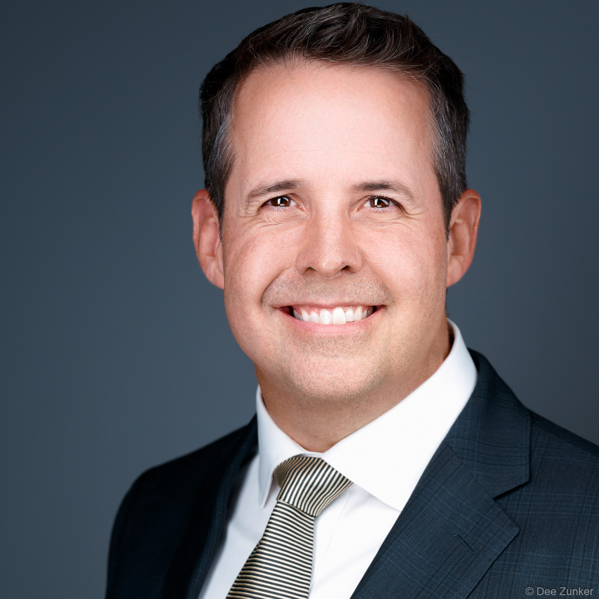 Smiling male executive in dark suit and striped tie, professional studio headshot with neutral grey backdrop, Bellaire Houston.