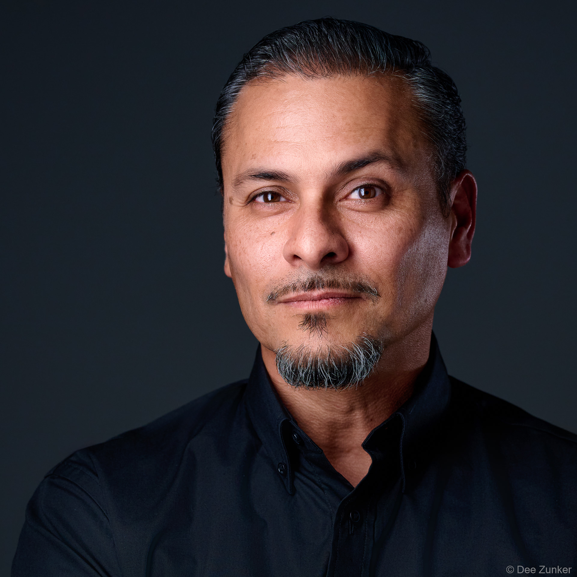 Male executive headshot on dark backdrop, slicked-back hair, goatee, black shirt, confident expression, studio lighting, Bellaire Houston.