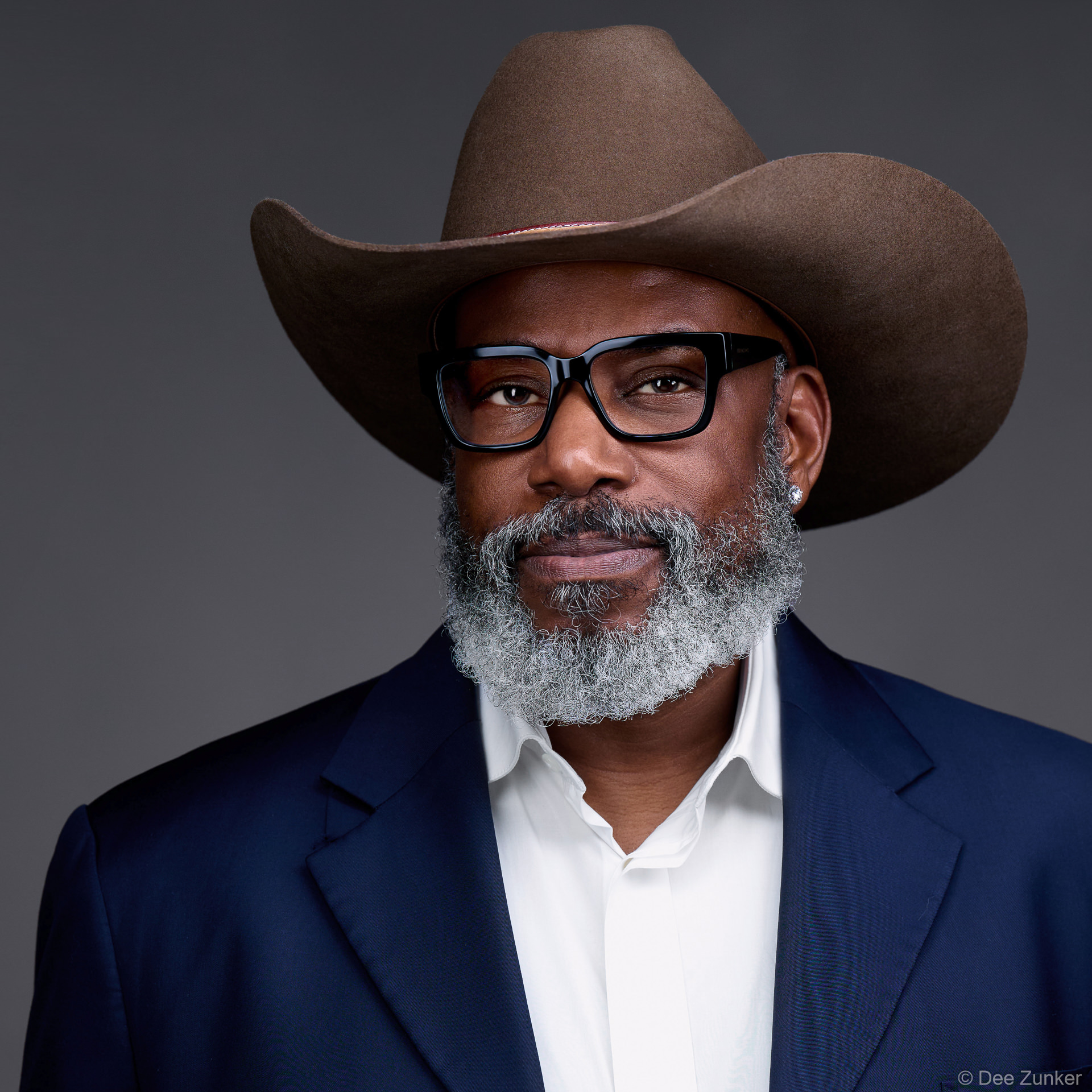 Male executive studio headshot in navy blazer and brown cowboy hat, grey beard, black glasses, neutral backdrop, Bellaire Houston.