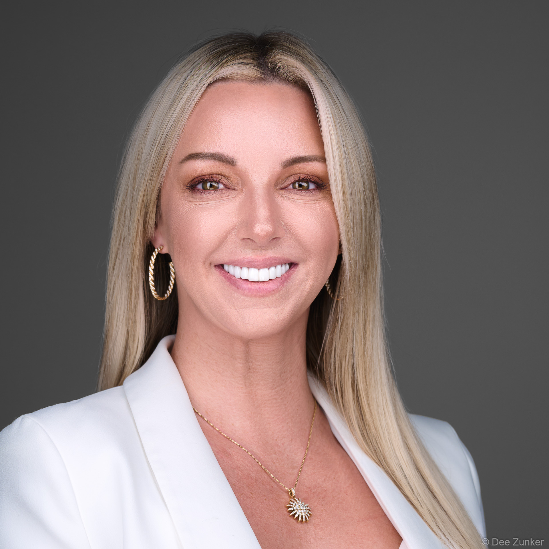 Woman in white blazer smiles confidently in a professional studio headshot, neutral grey backdrop, gold jewelry, Houston, Texas.