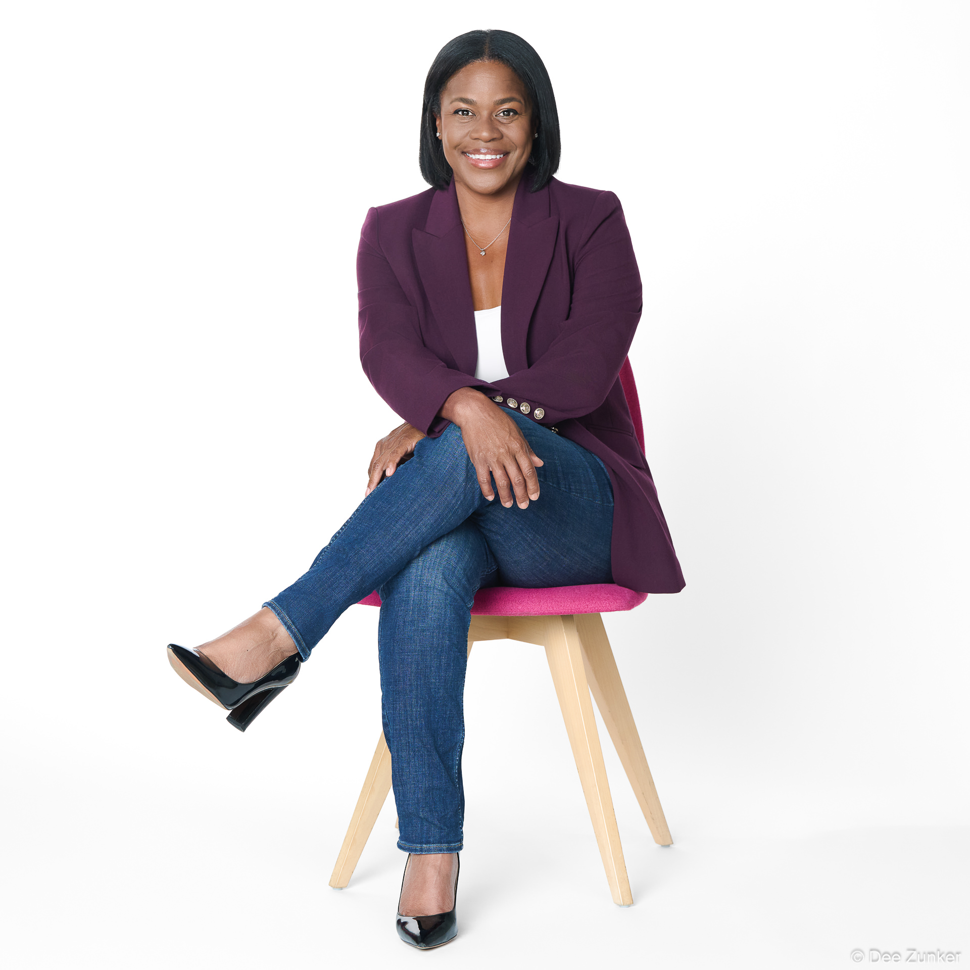 Full-body professional portrait of a woman in a purple blazer and jeans, seated cross-legged, smiling confidently against a white studio backdrop in Houston.