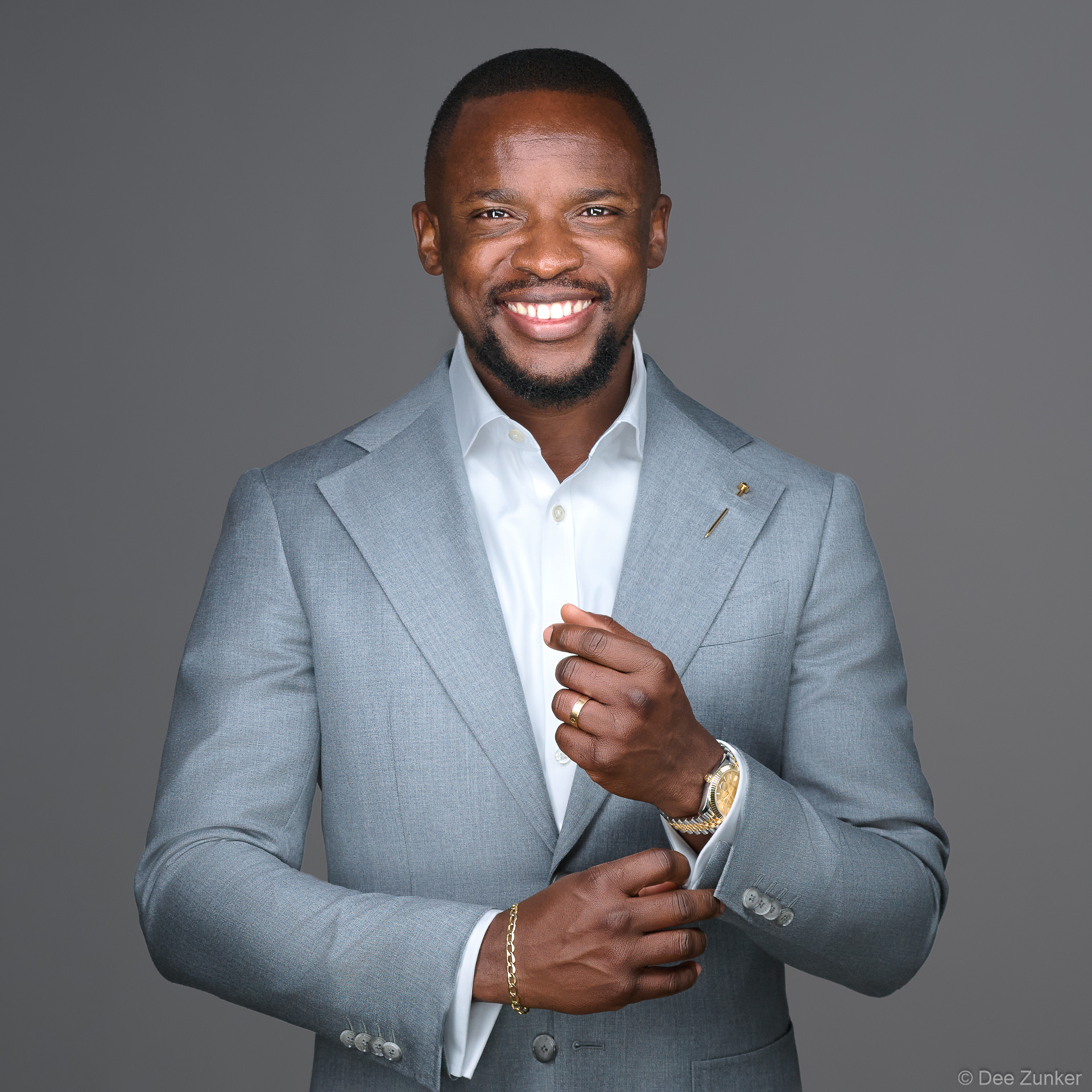 Smiling male executive in light gray suit adjusting cufflinks, full-body professional portrait, neutral gray backdrop, Houston studio session.