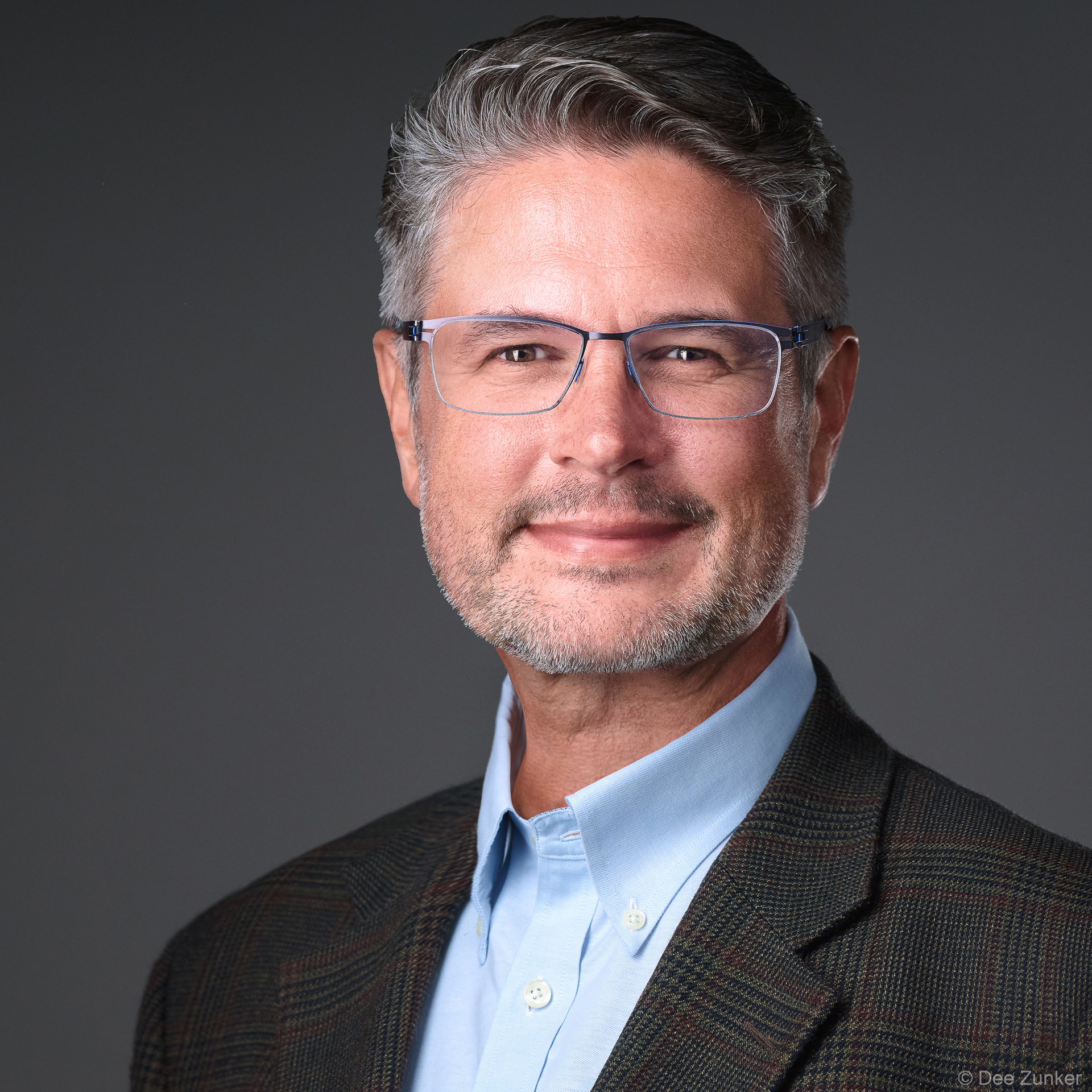 Male executive headshot in Houston studio, grey hair, glasses, plaid blazer, light blue shirt, warm smile, neutral grey backdrop.