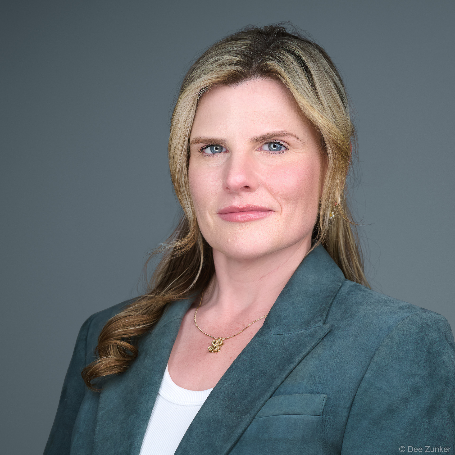 Woman in teal suede blazer and gold necklace poses for a serious executive studio headshot against a neutral gray backdrop in Houston.