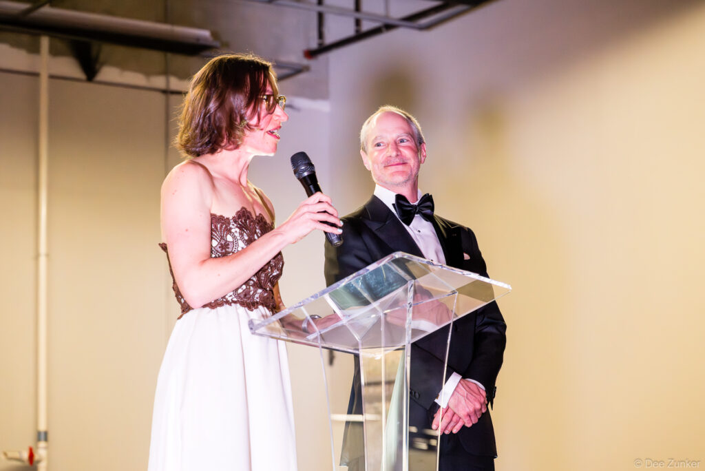 Woman in formal gown speaks at acrylic podium holding microphone beside man in tuxedo at AIA Houston Design Gala, The Shop Club.