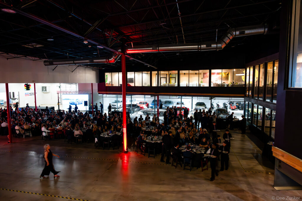 Elevated view of AIA Houston Design Gala dinner at The Shop Club, guests seated at round tables amid red-lit industrial venue with visible cars.