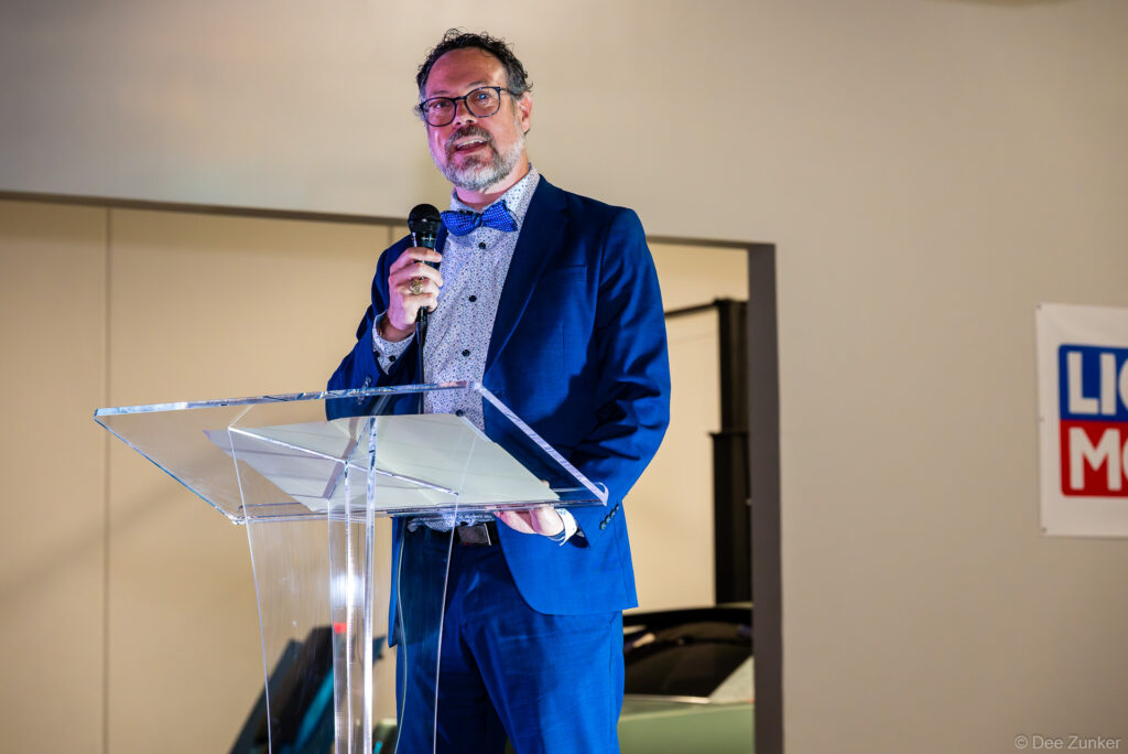Man in blue suit and bow tie speaks at clear acrylic podium during 40th Annual AIA Houston Design Gala at The Shop Club Houston.