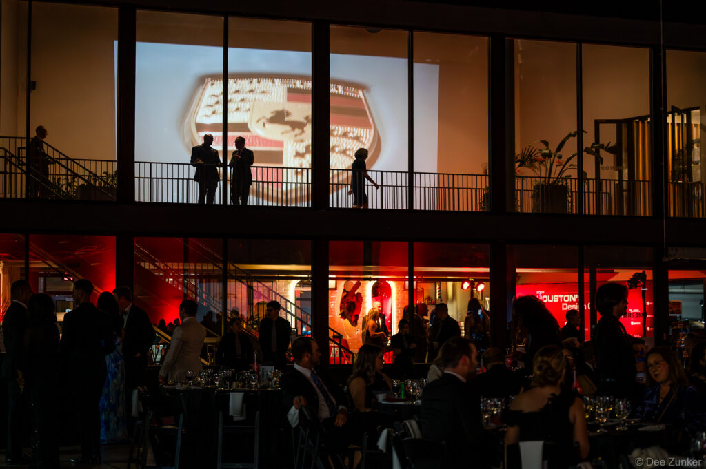 Silhouetted guests on a mezzanine balcony with a projected Porsche emblem above a gala crowd at The Shop Club Houston.