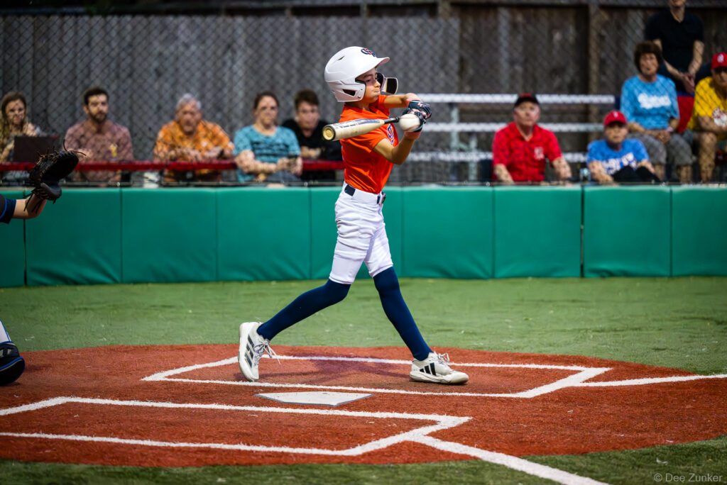 Young Astros batter in orange jersey follows through on a swing at home plate during a Bellaire Little League American League game in Bellaire TX.