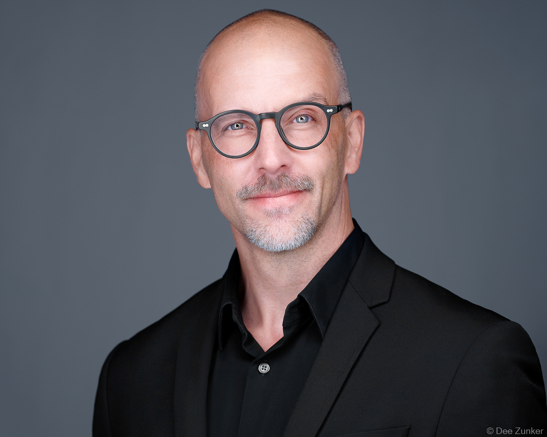 Bald man with round glasses and gray goatee smiling in black blazer, delivered as sponsor activation headshot booth attendee 