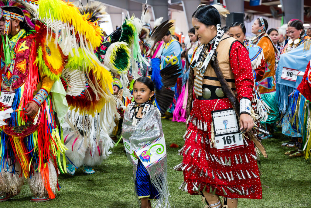 Native American dancers in traditional regalia including a young girl and woman in red jingle dress at Alabama-Coushatta Annual Powwow Grand Entry Texas