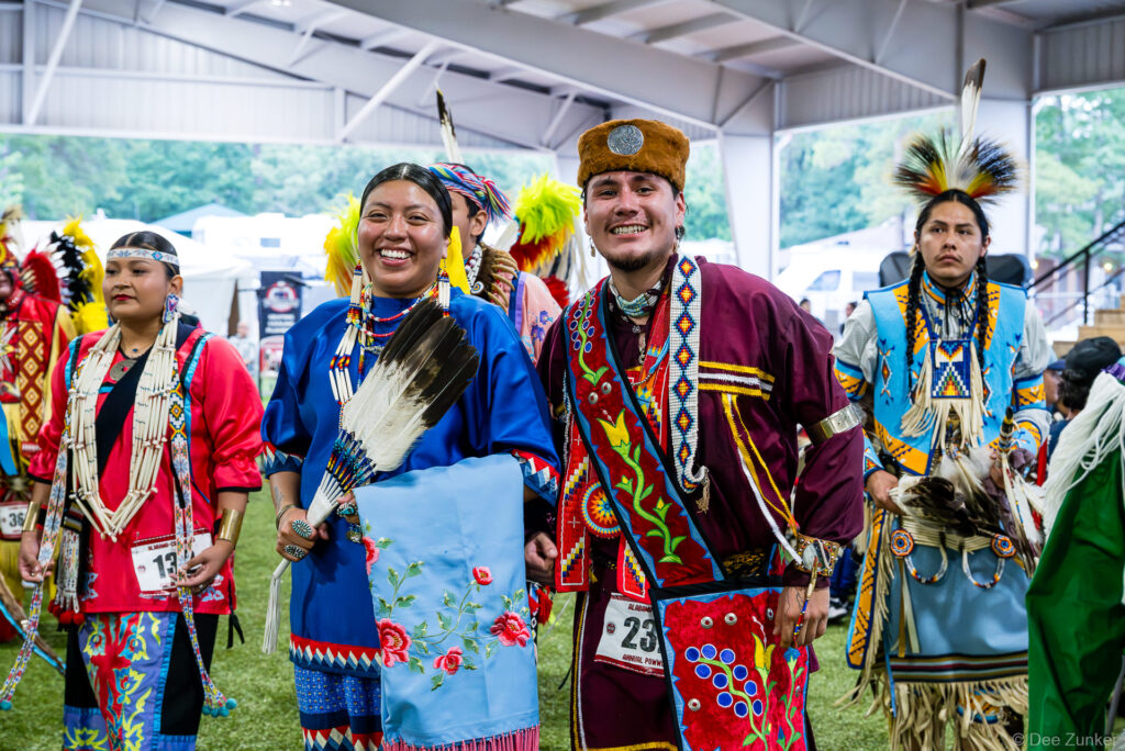 Native American dancers in traditional regalia smiling at the Alabama-Coushatta 56th Annual Powwow in Texas, June 2025.