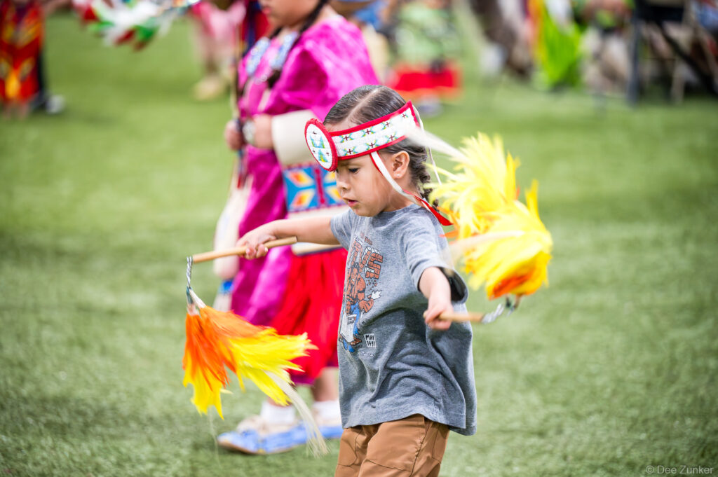 Young Native American child dancing at Alabama-Coushatta 56th Annual Powwow, wearing beaded headband and holding colorful feathered fans, Texas
