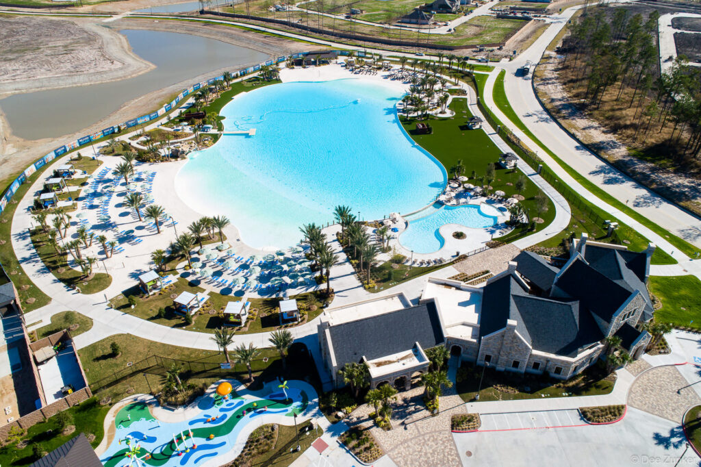Drone aerial photo of a large crystal lagoon pool with lounge areas, palm trees, splash pad, and clubhouse in a Houston residential community.