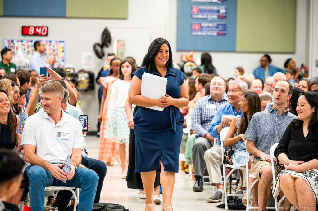 Principal or teacher in navy dress walks aisle holding papers at Horn Elementary 5th grade graduation ceremony in Bellaire TX