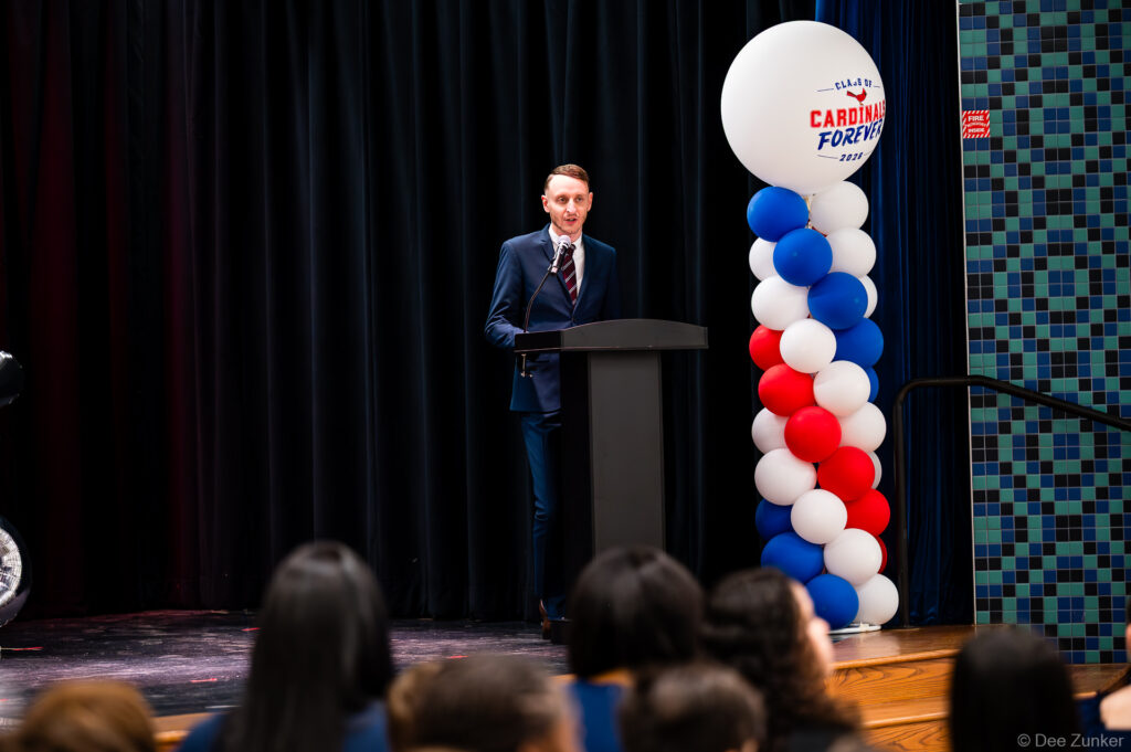 Speaker at podium addresses Horn Elementary 5th grade graduation ceremony in Bellaire TX beside red white and blue balloon column