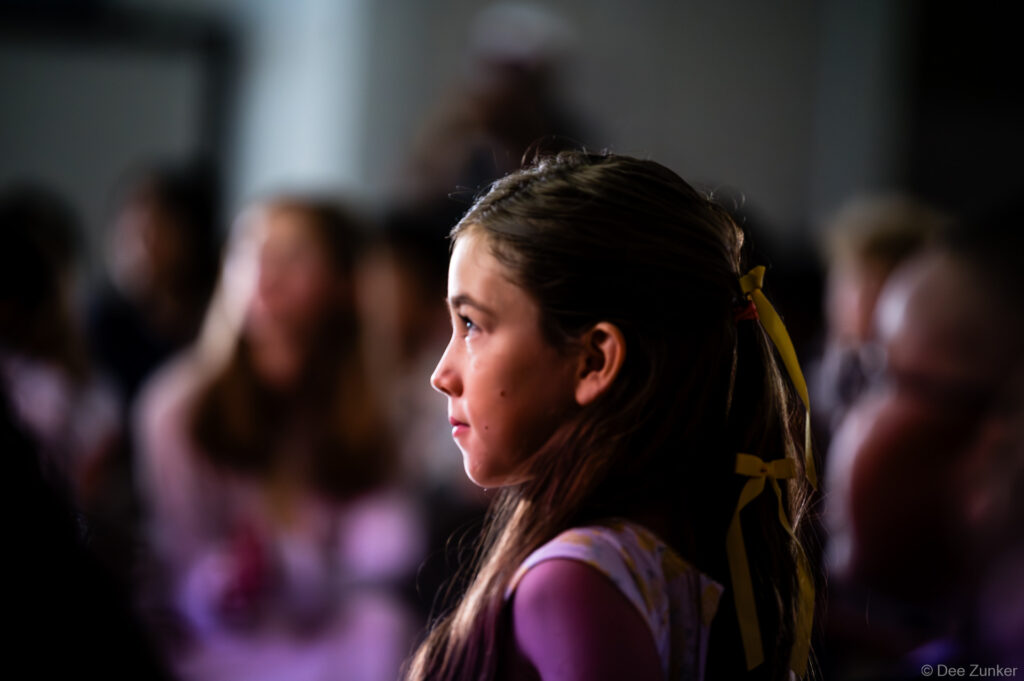 Young girl in profile with yellow ribbon hair bows watches attentively during Horn Elementary 5th grade graduation ceremony in Bellaire TX