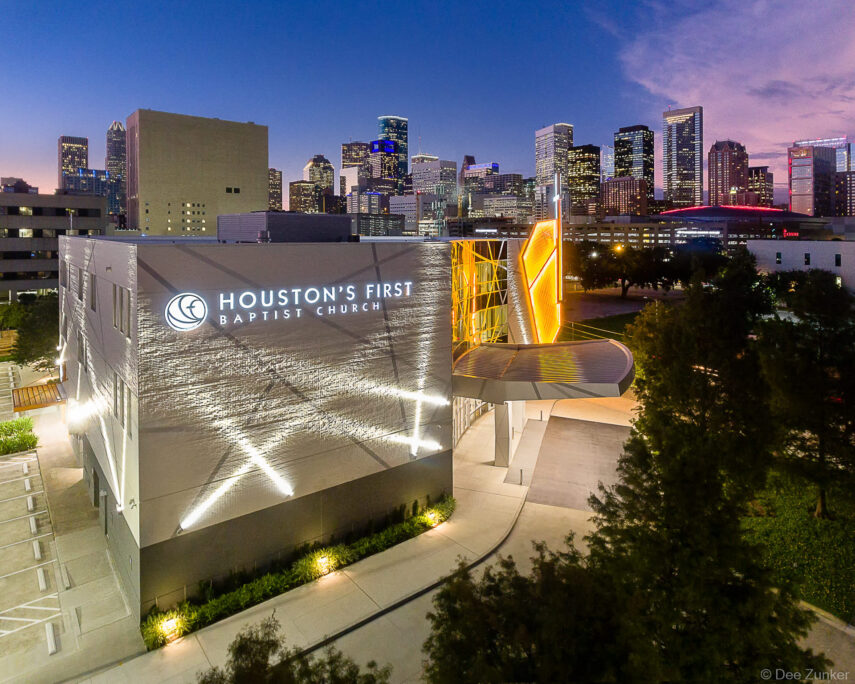 Aerial twilight exterior of Houston's First Baptist Church downtown campus with illuminated facade and Houston skyline in the background.