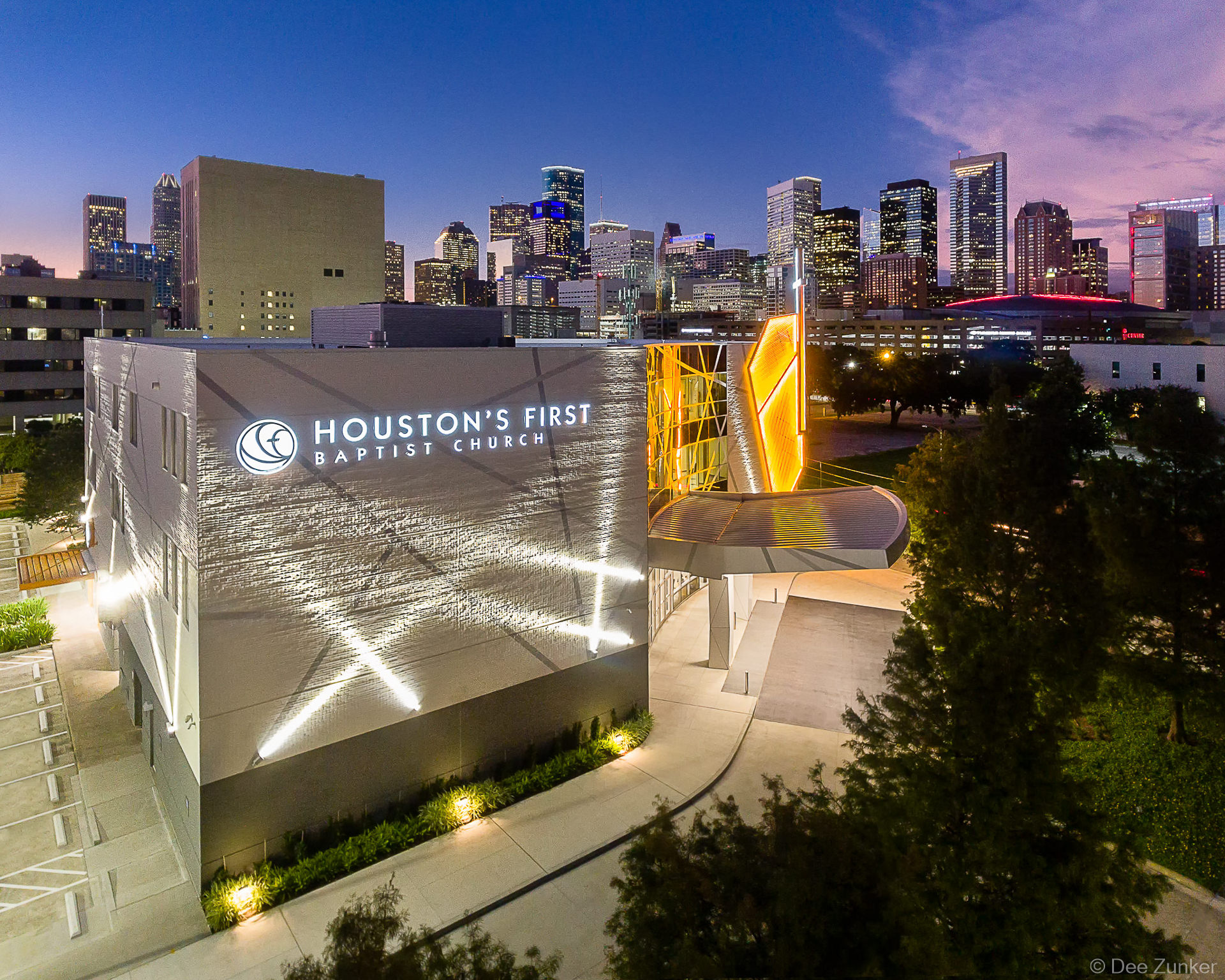 Aerial twilight exterior of Houston's First Baptist Church downtown campus with illuminated facade and Houston skyline in the background.