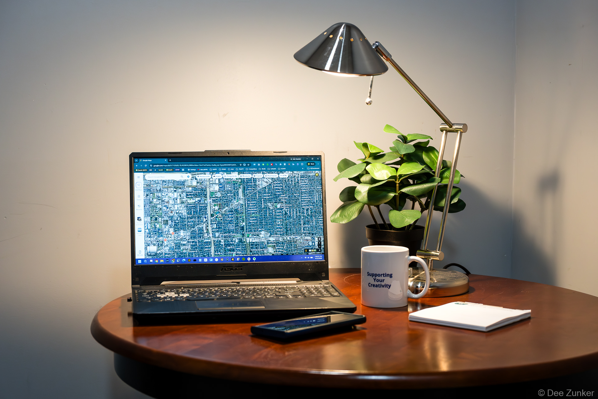 ASUS laptop displaying Google Maps satellite view on a wood desk with lamp, plant, and Supporting Your Creativity coffee mug.