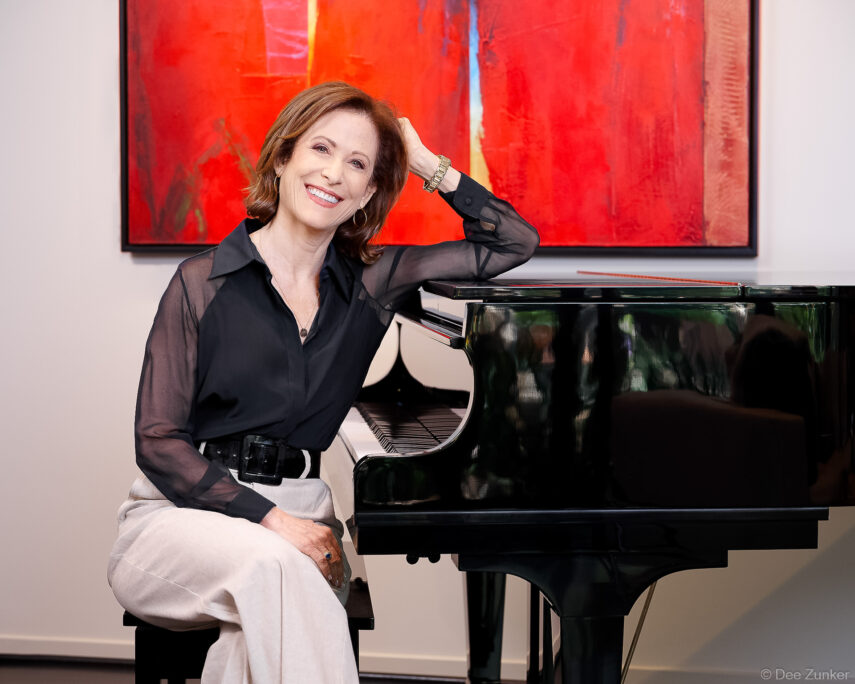 Woman photographer poses beside a black grand piano in a studio with vibrant red abstract art, Houston professional branding portrait.