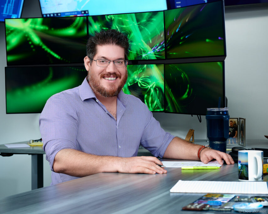 Smiling male professional in blue shirt seated at standing desk with six green-lit monitors, corporate headshot session, Houston TX.