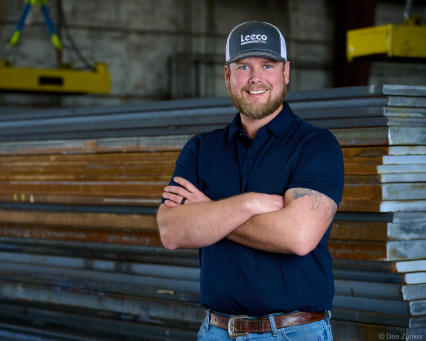 Smiling bearded man in navy polo and branded cap stands arms crossed before stacked steel plates in an industrial facility, corporate staff headshot.