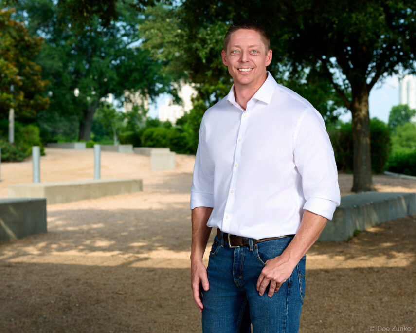 Three-quarter environmental portrait of a smiling male professional in white shirt and jeans outdoors at a Houston park, corporate headshot